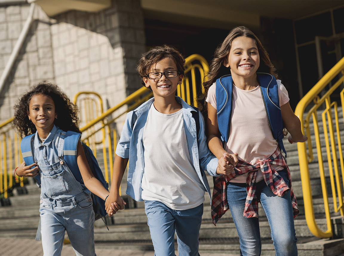 Three happy, young school children wearing backpacks, holding hands and walking