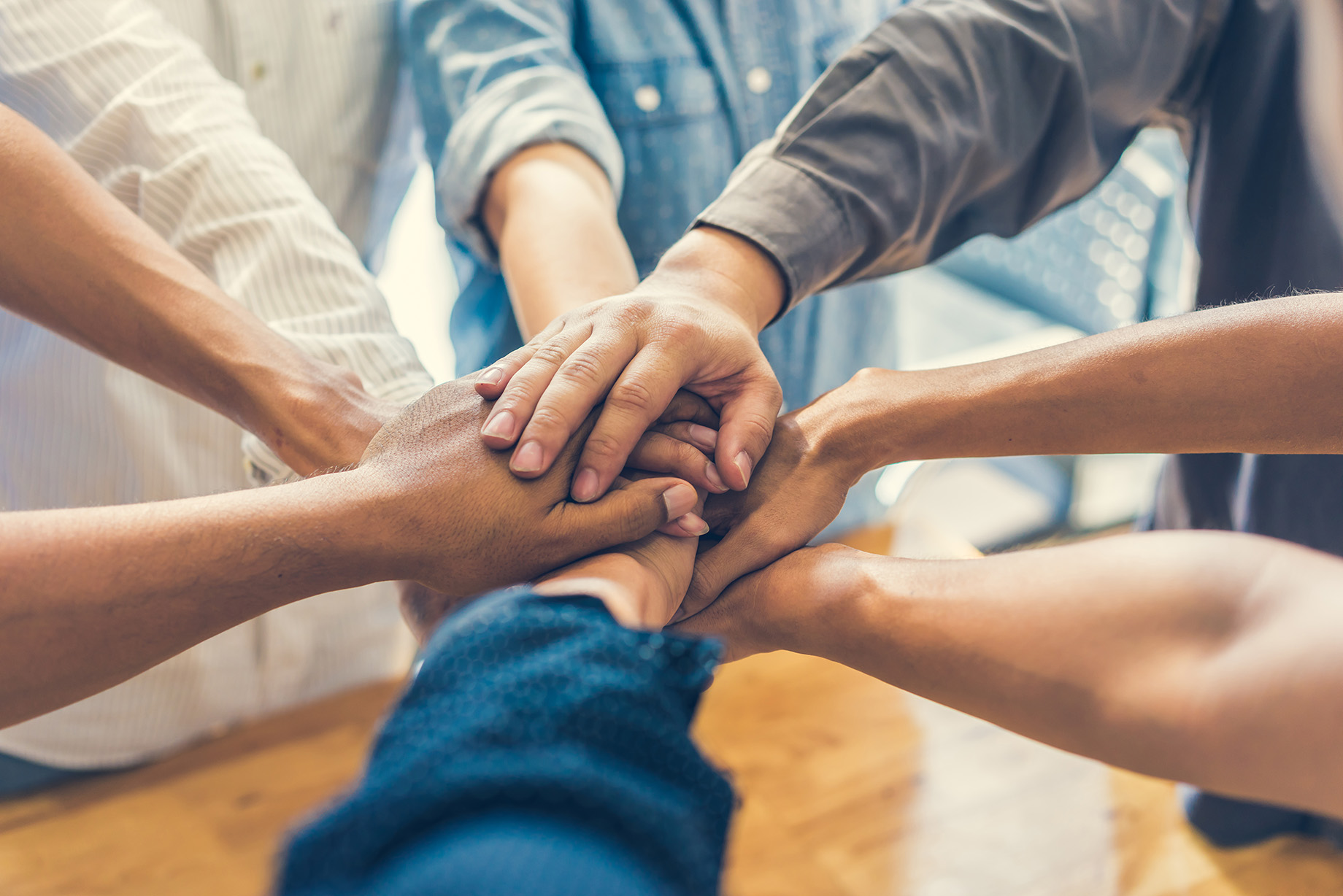 Several people from diverse backgrounds place their hands together in a stack, forming a circle to symbolize teamwork and collaboration in an indoor setting.
