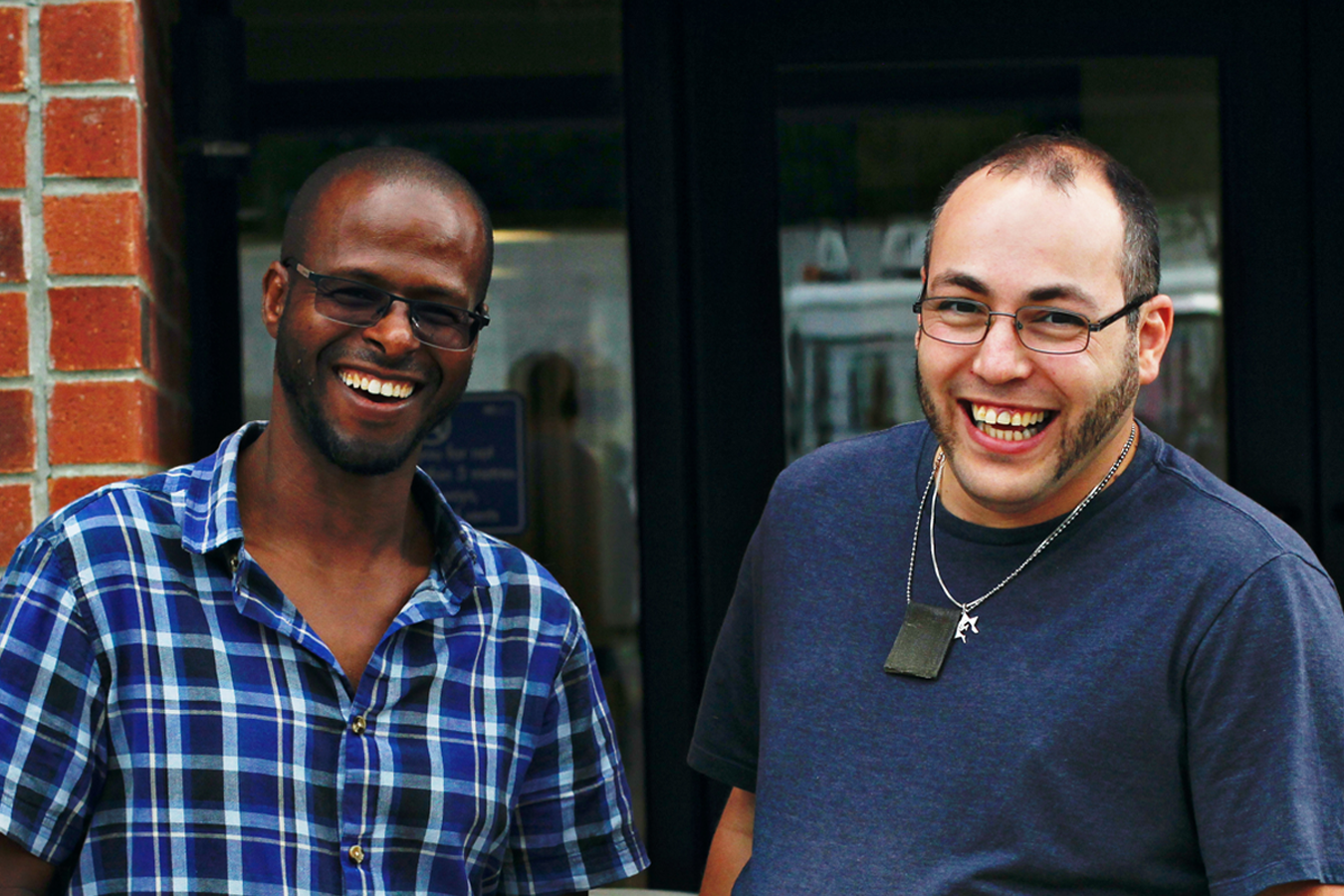 Two individuals standing outside a building entrance, one wearing a blue plaid shirt and the other in a dark t-shirt.