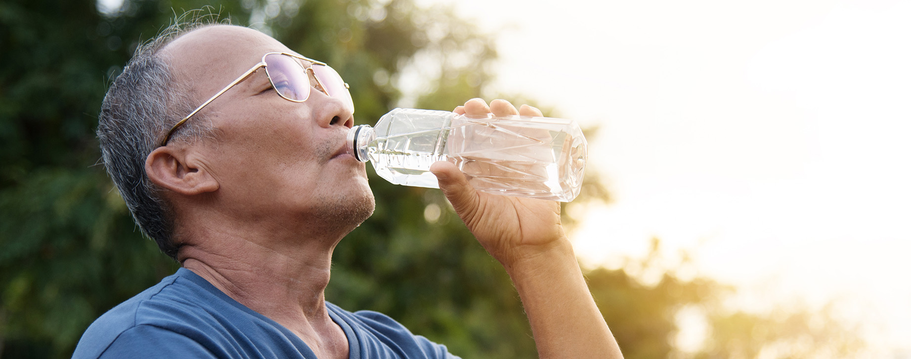 Senior wearing glasses and a blue t-shirt drinking water outdoors with the sun shining in the background.