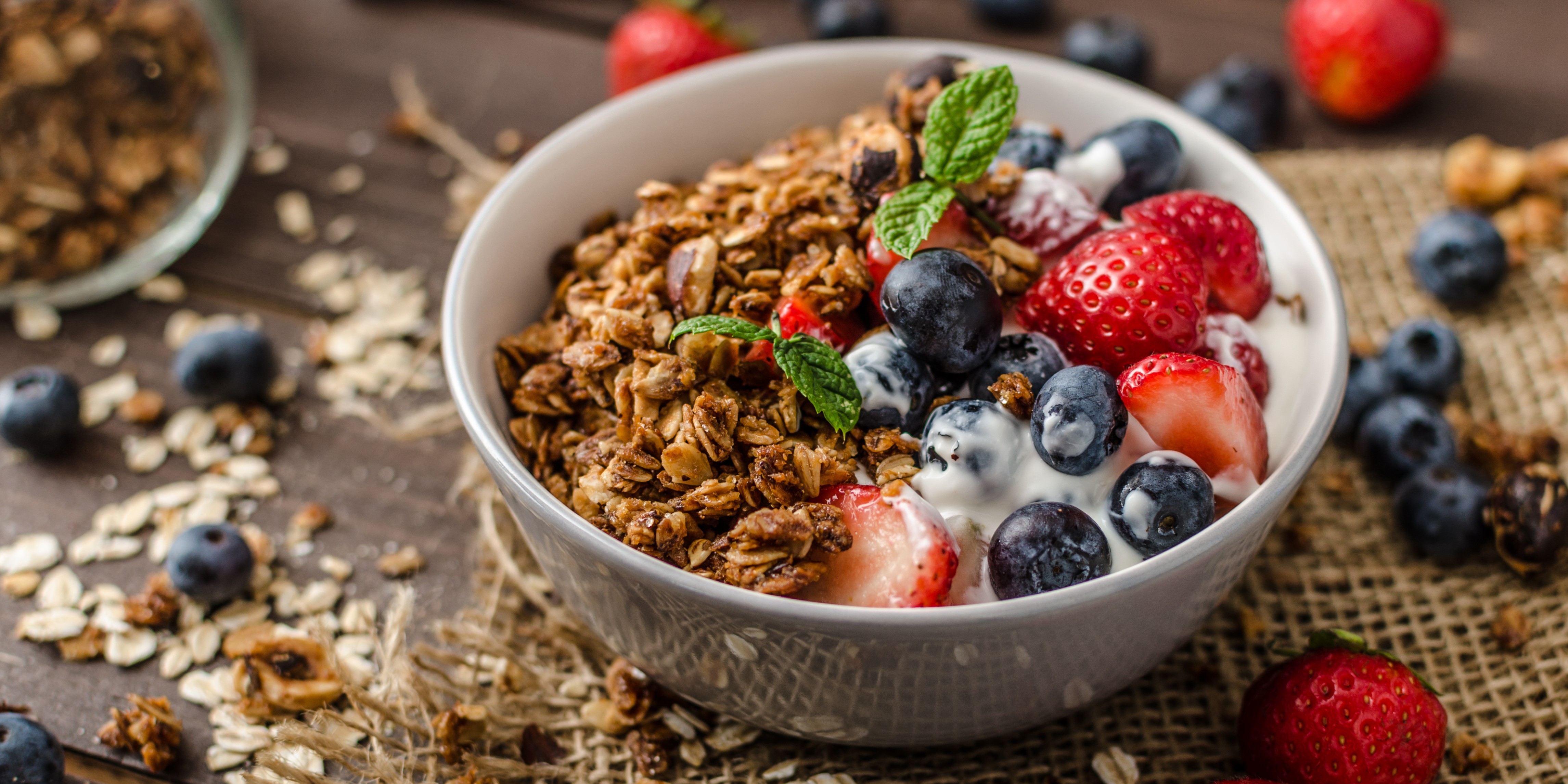 Bowl of homemade granola with yogurt, fresh strawberries, blueberries, and mint on a rustic tabletop.