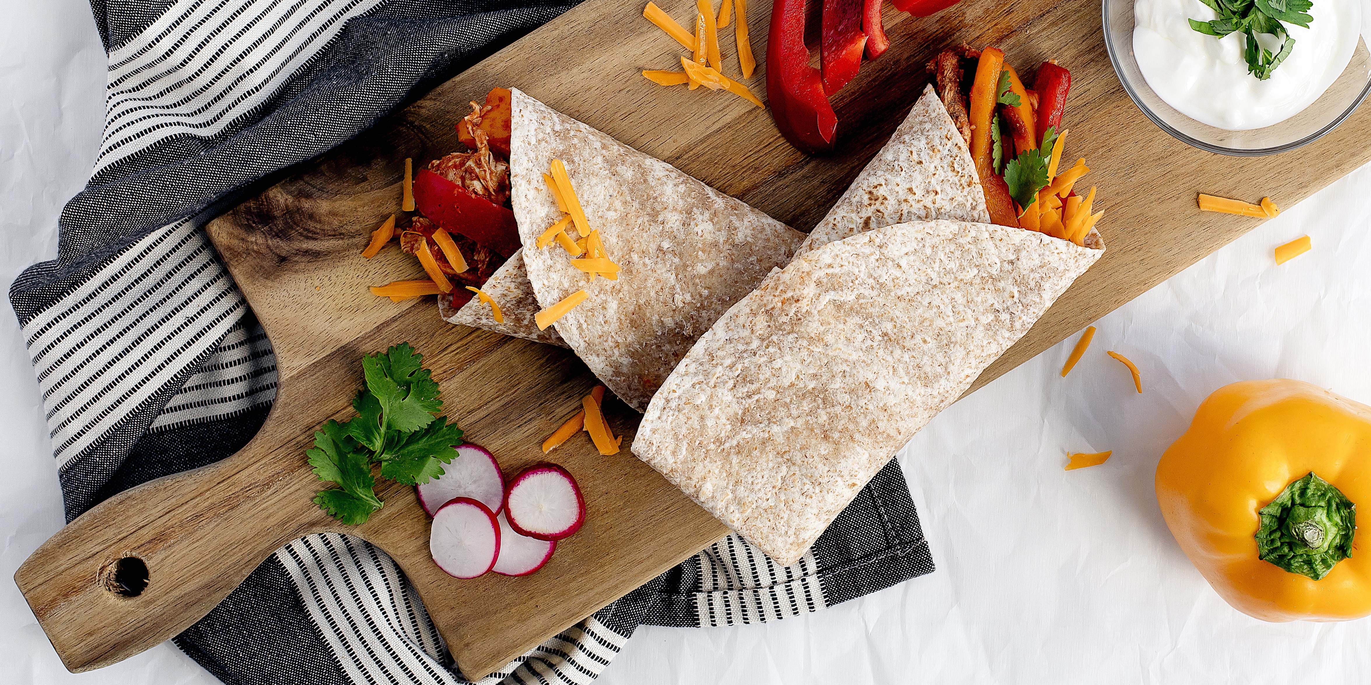 Whole‑wheat chicken fajita wraps with peppers and shredded cheese on a wooden cutting board, garnished with radishes and herbs.