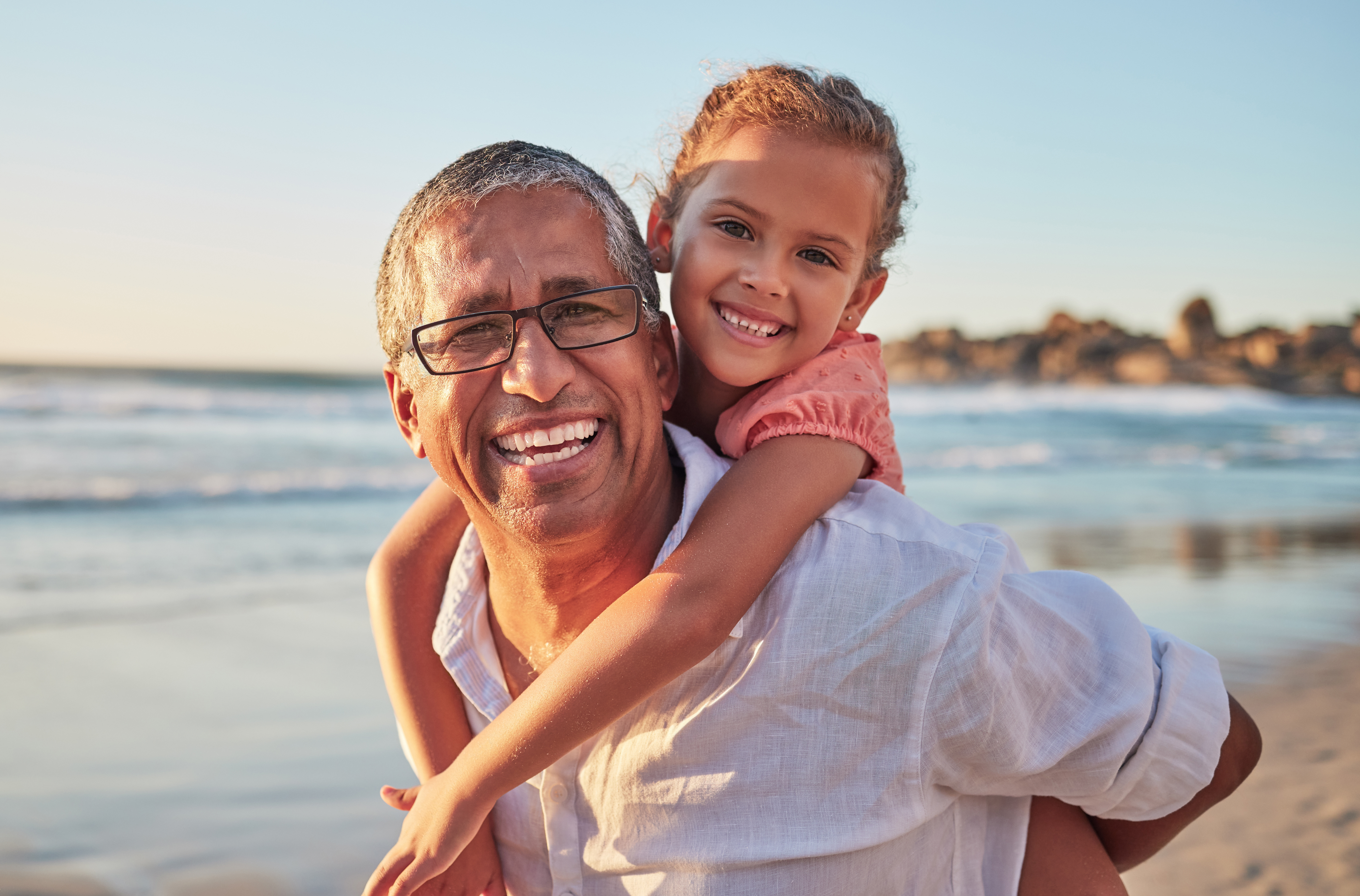 Older adult wearing glasses giving a young child a piggyback ride on a beach, with ocean waves and rocky shoreline in the background.