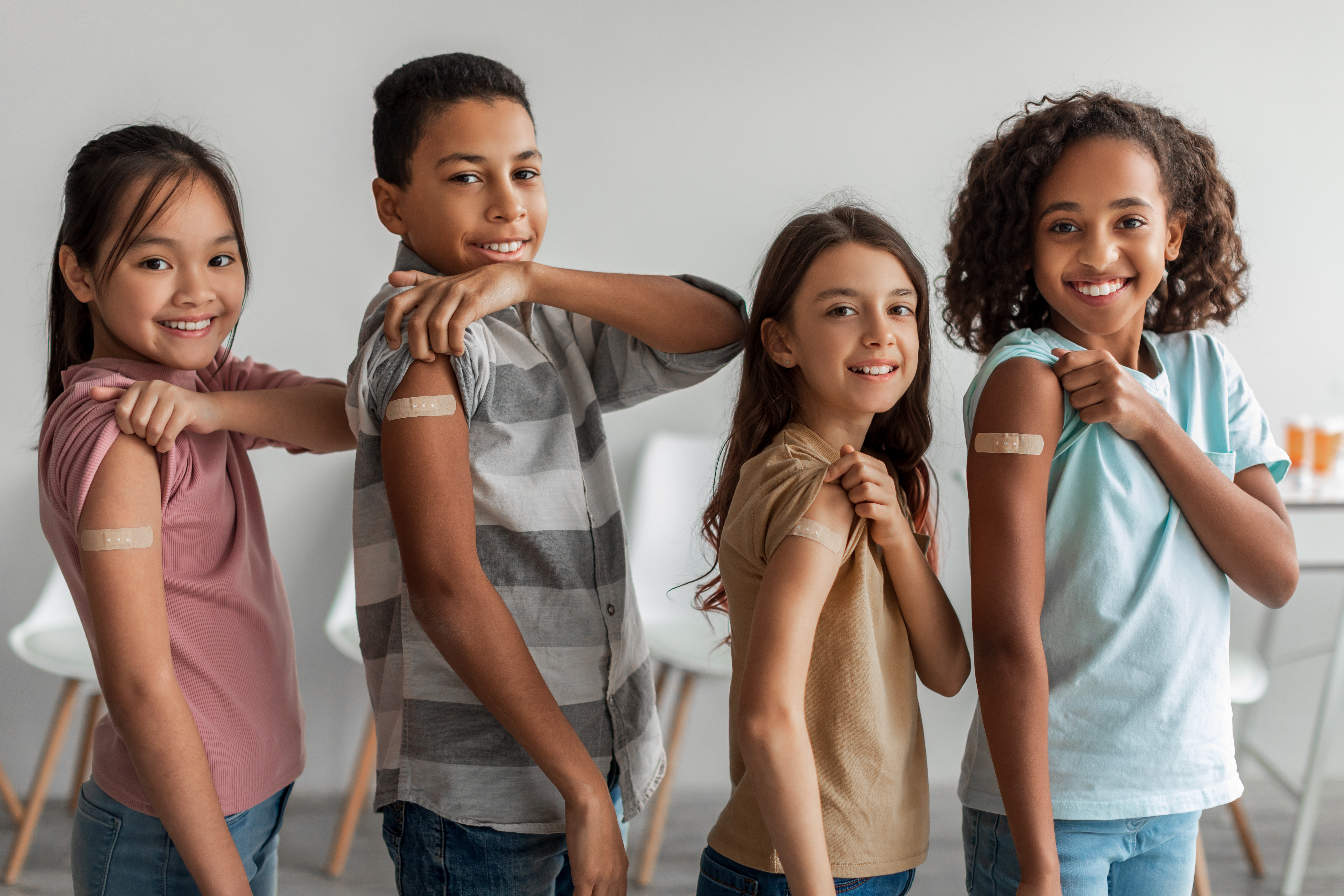 Four children of different ages and genders smiling and rolling up their sleeves to show a band aid on their upper arms indicating a received vaccination.