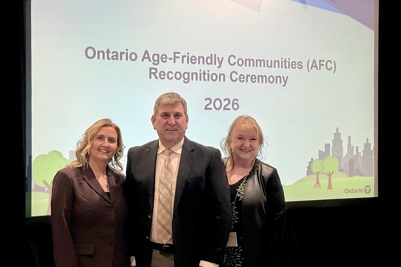 Three individuals smiling and posing in front of an Ontario Age-Friendly Communities (AFC) poster