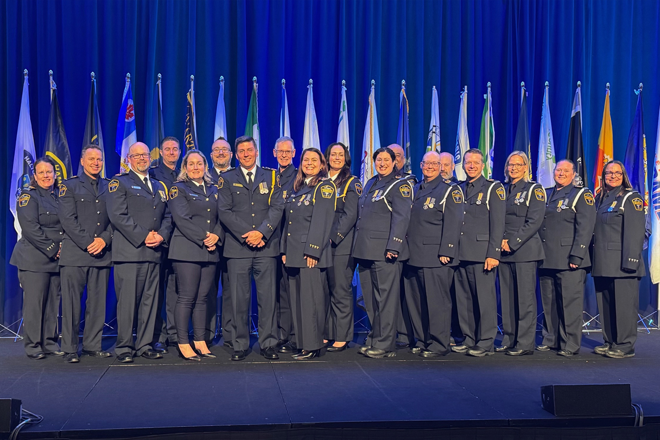 Group portrait of paramedics who received the Governor General's Emergency Medical Services (EMS) Exemplary Service Medal. The recipients stand on a stage with medals displayed on their uniforms, with a row of municipal and organizational flags and a blue-lit backdrop behind them.