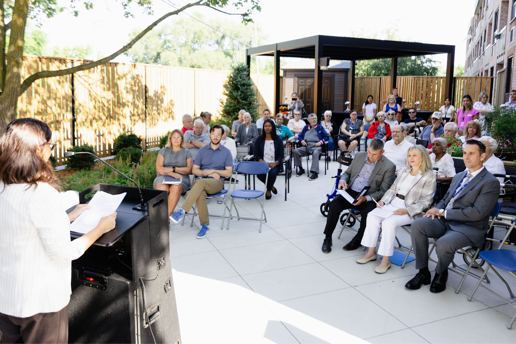 Speaker addresses attendees at an outdoor gathering to celebrate the renovation of the Keswick Adult Day Program's backyard patio and garden