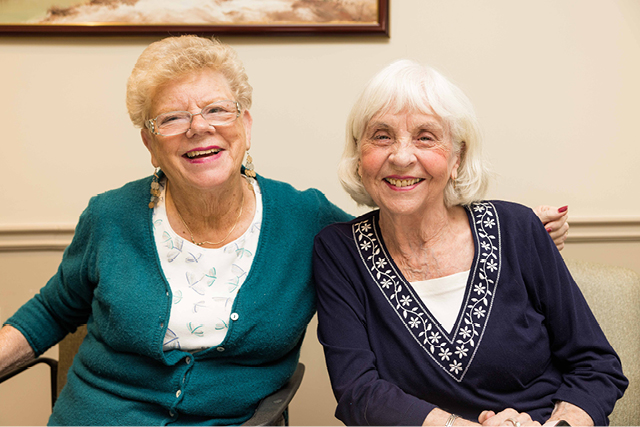 Two elderly women sitting side by side indoors, facing the camera and smiling.