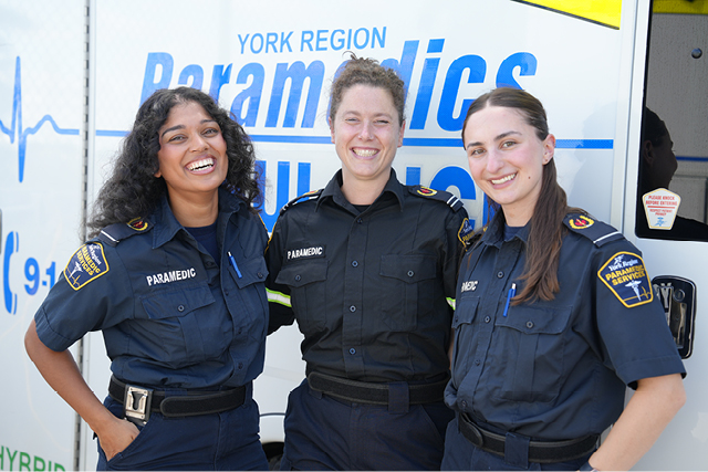 Three uniformed paramedics standing together in front of a York Region Paramedics ambulance, with visible shoulder patches and badges on their shirts.