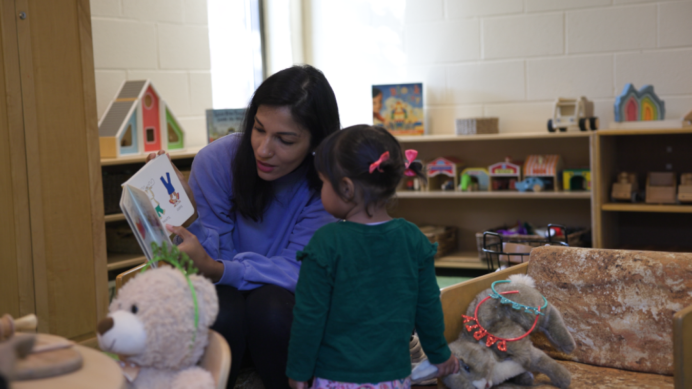A caregiver reads a picture book to a young child in a playroom filled with toys and stuffed animals.