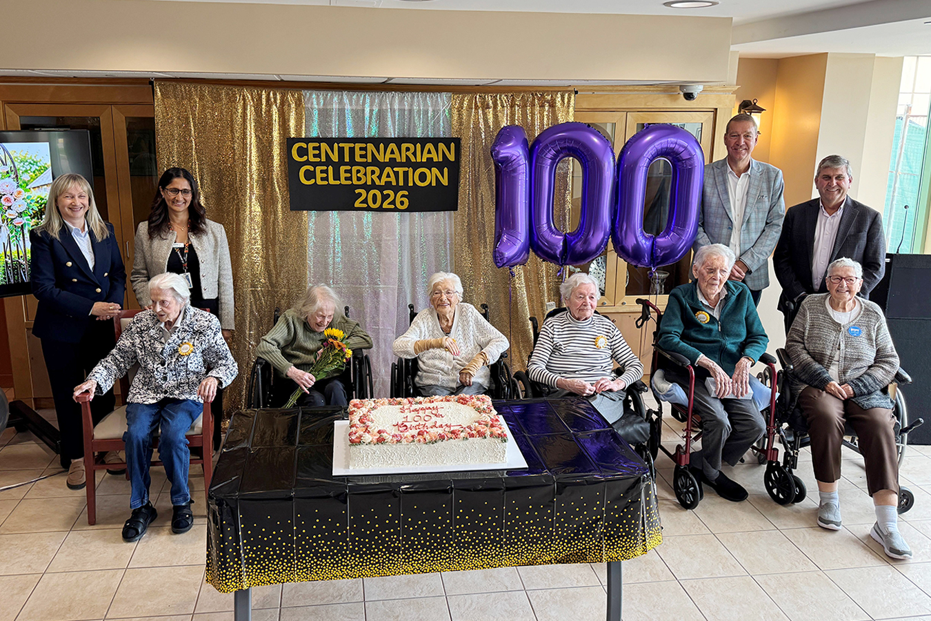 Centenarians gather around a birthday cake beneath a “Centenarian Celebration 2026” sign and purple “100” balloons during a milestone celebration.