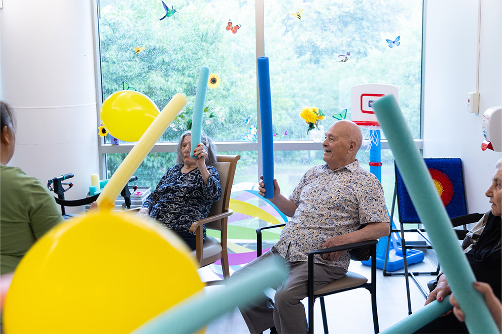 A group of seniors participate in a fun, active recreation program, using pool noodles and balloons to engage in a cooperative exercise activity.