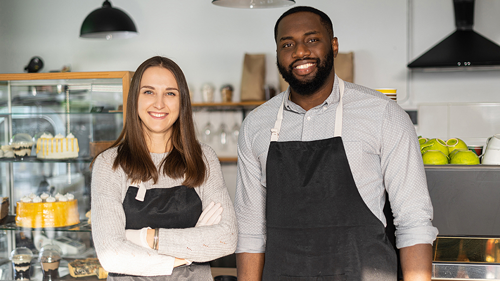 A man and a women wearing uniforms standing inside a bakery smiling