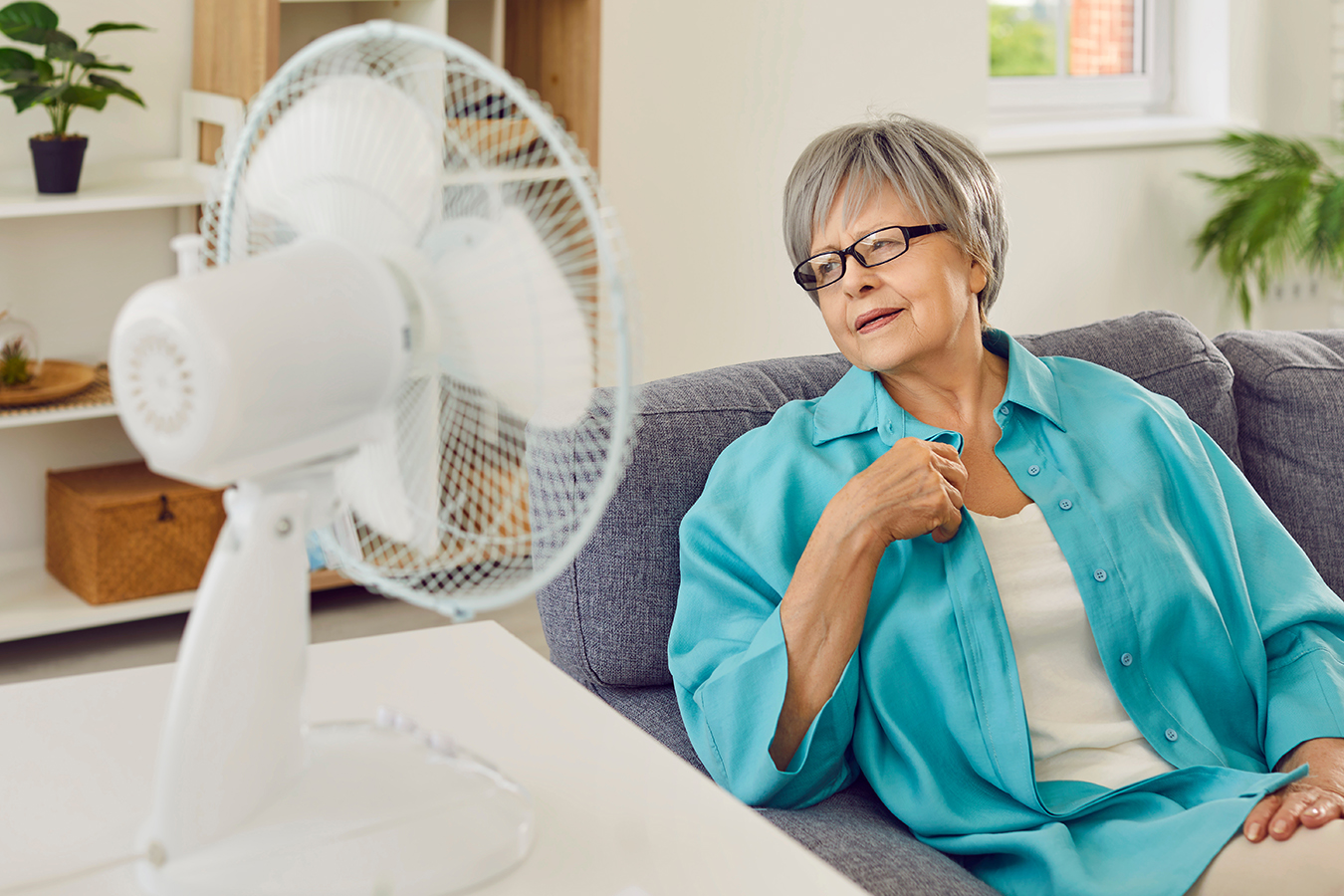 Older person wearing glasses and a blue button-down shirt sitting by a fan and looking away