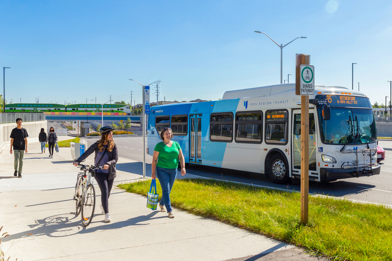 people walking and cycling on a shared pathway near a transit bus and a GO train