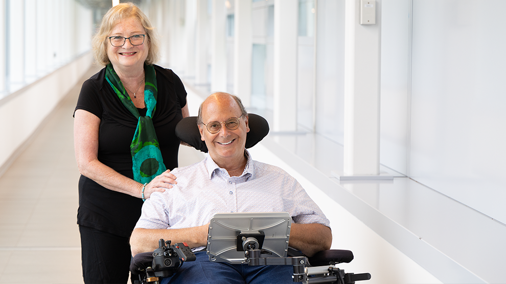 Current committee members Kathleen standing and smiling beside Steve seated and smiling in his power wheelchair