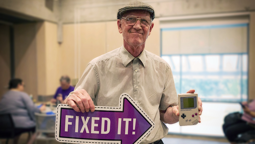 an older man holding a sign that reads fixed it in one hand and the other hand holding an electronic game.