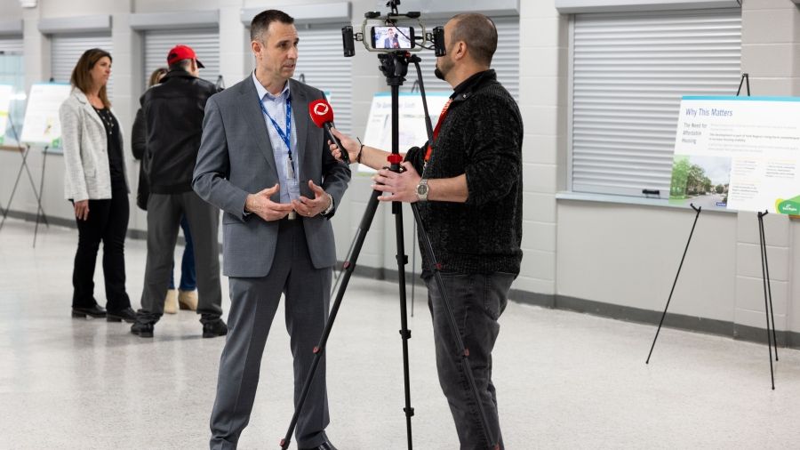 A person is being interviewed on camera at The Queensway South community open house, with display boards and attendees in the background.