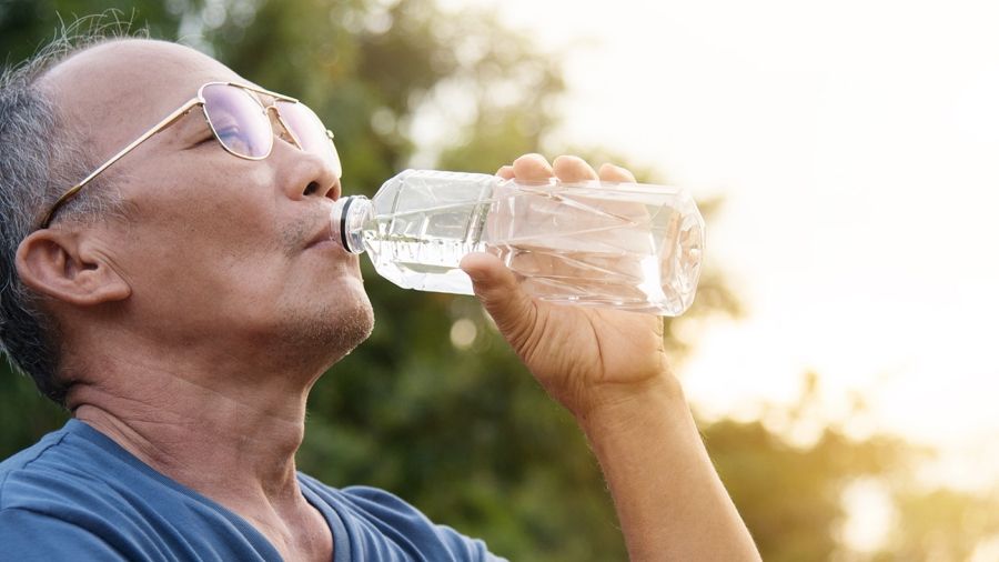 Senior wearing glasses and a blue t-shirt drinking water outdoors with the sun shining in the background.
