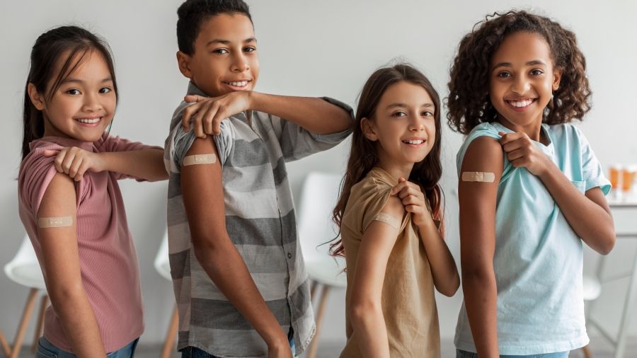 Four children of different ages and genders smiling and rolling up their sleeves to show a band aid on their upper arms indicating a received vaccination.