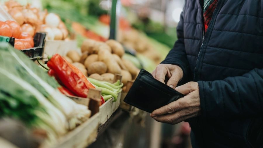 Person holding a wallet while standing beside a display of fresh vegetables at a grocery store.
