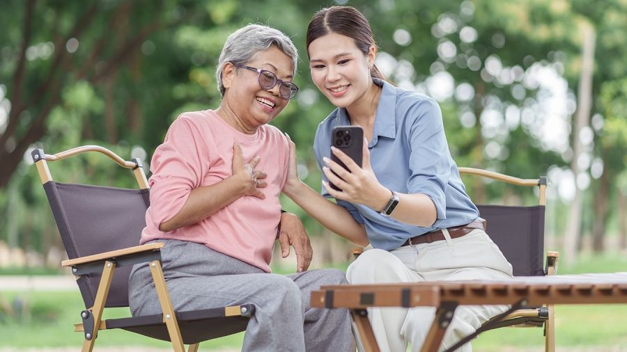 An older adult and a younger adult sitting outside, looking at a smart phone together and smiling.