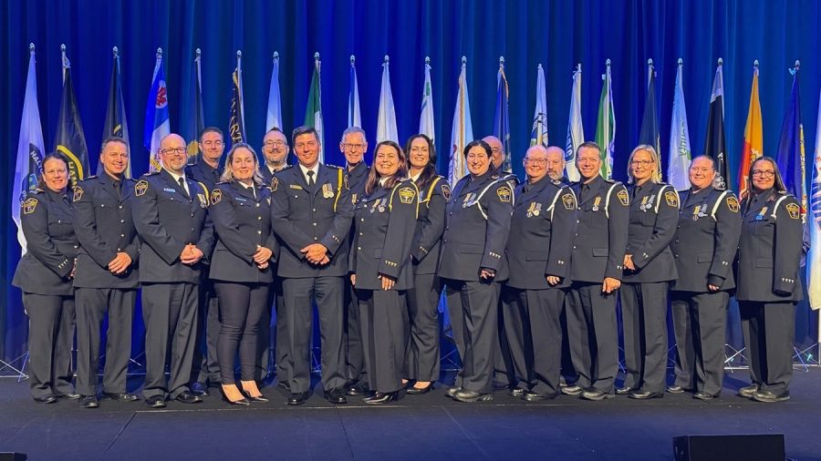 Group portrait of paramedics who received the Governor General's Emergency Medical Services (EMS) Exemplary Service Medal. The recipients stand on a stage with medals displayed on their uniforms, with a row of municipal and organizational flags and a blue-lit backdrop behind them.