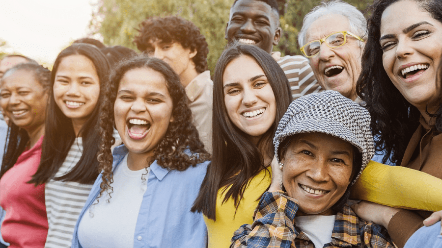 Multicultural and multi-generational group of nine smiling people; trees and sunshine are in the background