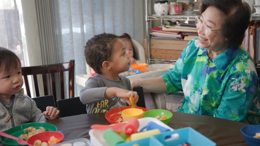 Adult engaging with young children at a dining table during a shared meal and learning activity.