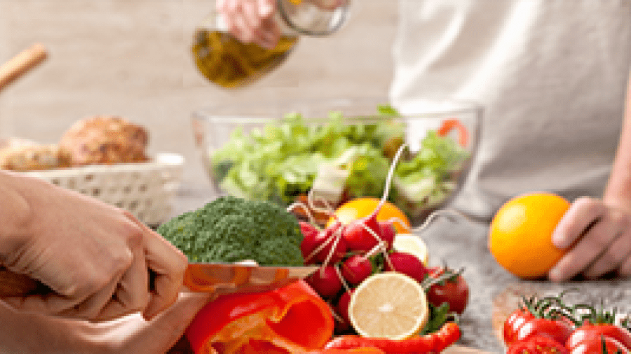 A woman slices vegetables while a man pours dressing on a green salad.
