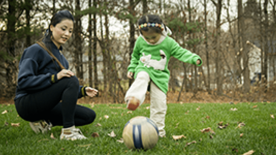 A young girl kicking a soccer ball while her mother watches
