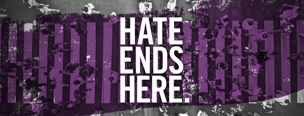 A black and white aerial image of people crossing a busy crosswalk with bold white stripes. Centered is a transparent purple brush stroke with bold white text reading, ‘Hate Ends Here’