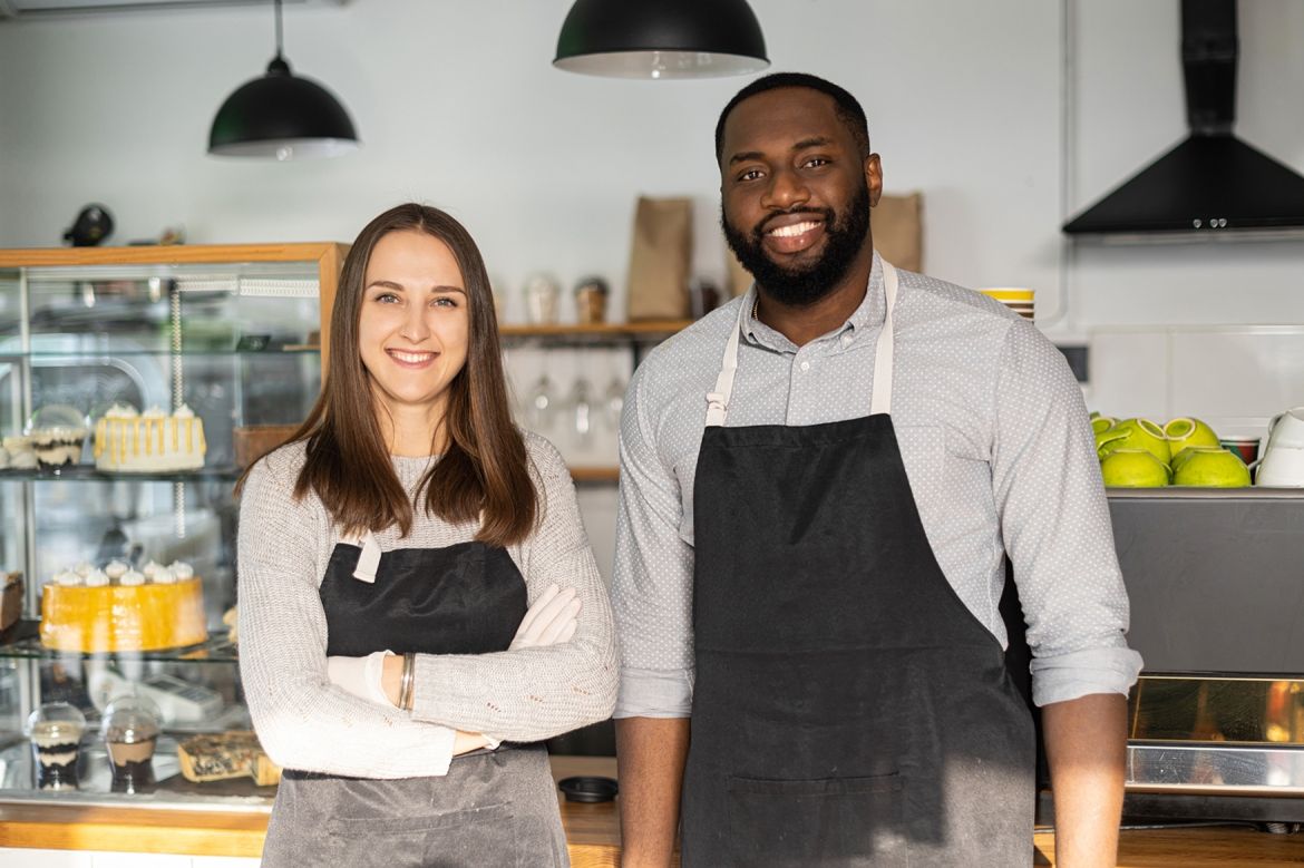 A man and a women wearing uniforms standing inside a bakery smiling
