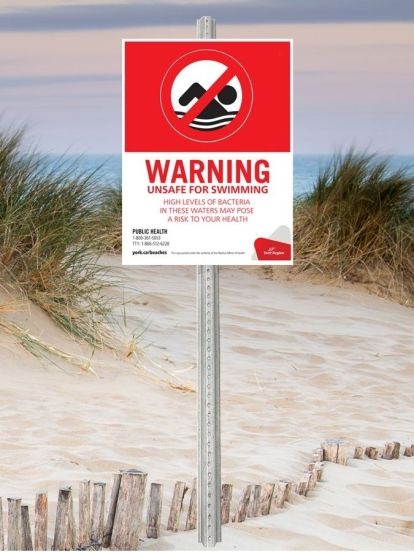 York Region Beach Water Warning Sign with an icon of a person swimming crossed out on a red background. On a white background, the sign states: "Warning, unsafe for swimming. High levels of bacteria in these waters may pose a risk to your health. Public Health: 1-800- 361-5653; TTY: 1-866-512-6228; york.ca/beaches"