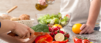 A woman slices vegetables while a man pours dressing on a green salad.